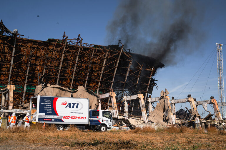 Damaged building with smoke billowing from it, an ATI restoration truck parked nearby, and workers in protective gear.