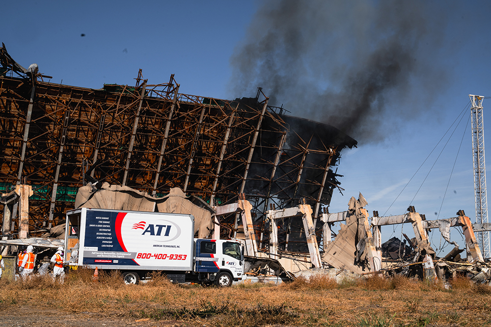 Damaged building with smoke billowing from it, an ATI restoration truck parked nearby, and workers in protective gear.