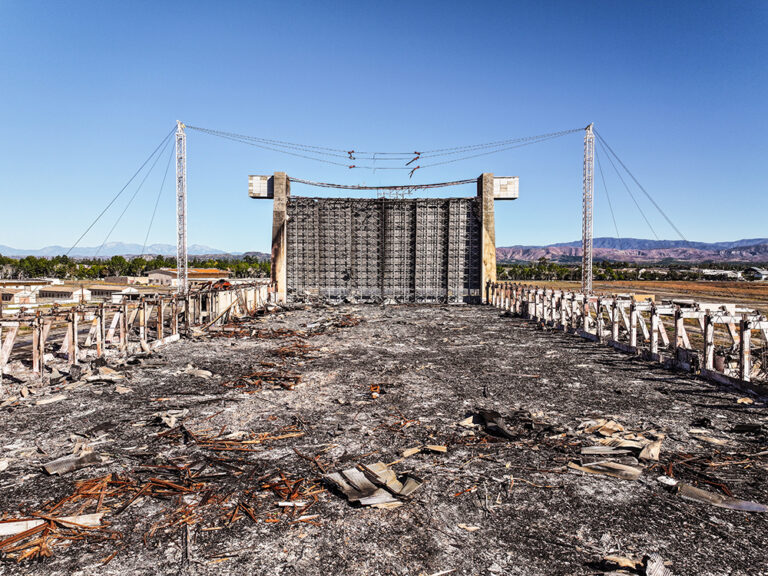 Burned remains of a large structure with a tall screen in the background.