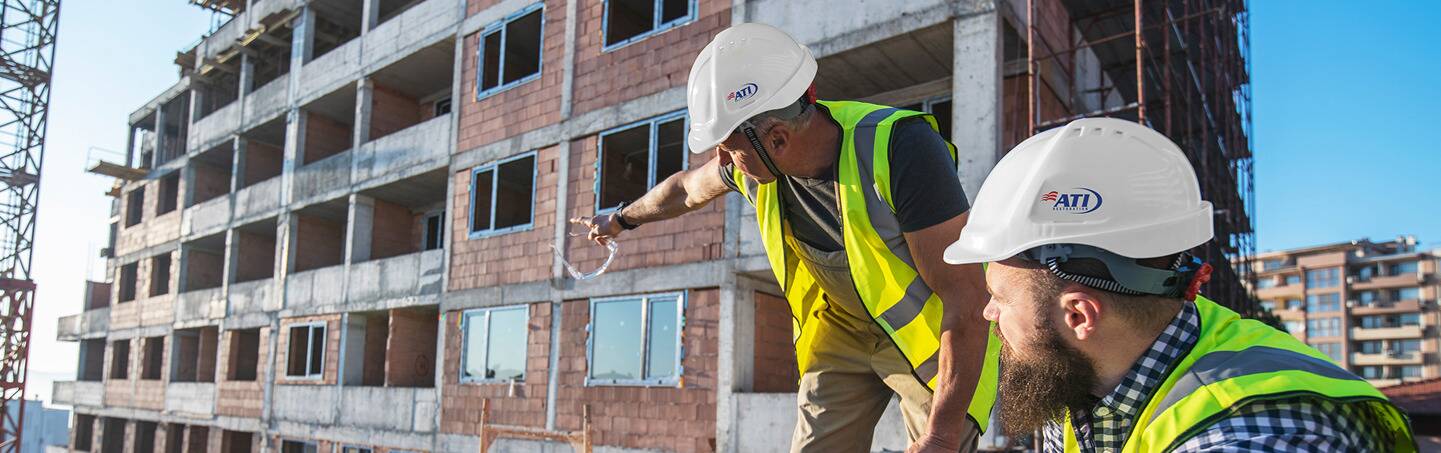 Two construction workers in hard hats and safety vests look at a building under construction.