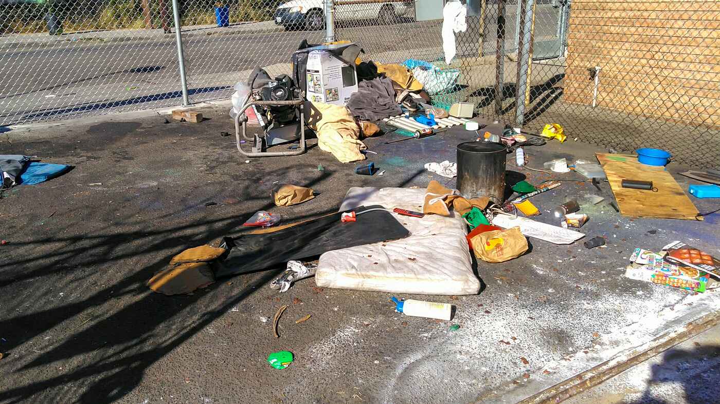 A mattress and various discarded items, including clothing, trash, and a small generator, are strewn on the ground outside a chain-link fence.