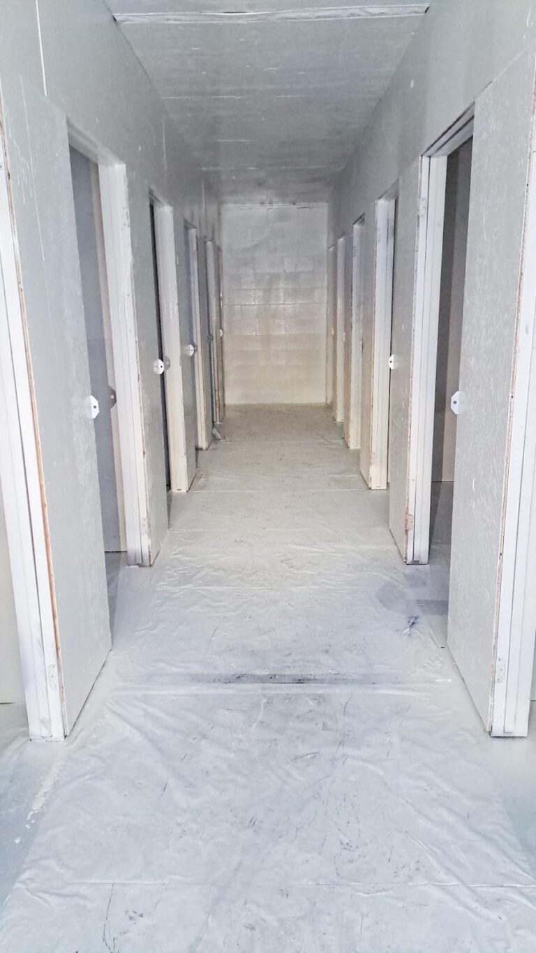 Long hallway with many white doors and a white floor covered in plastic sheeting.