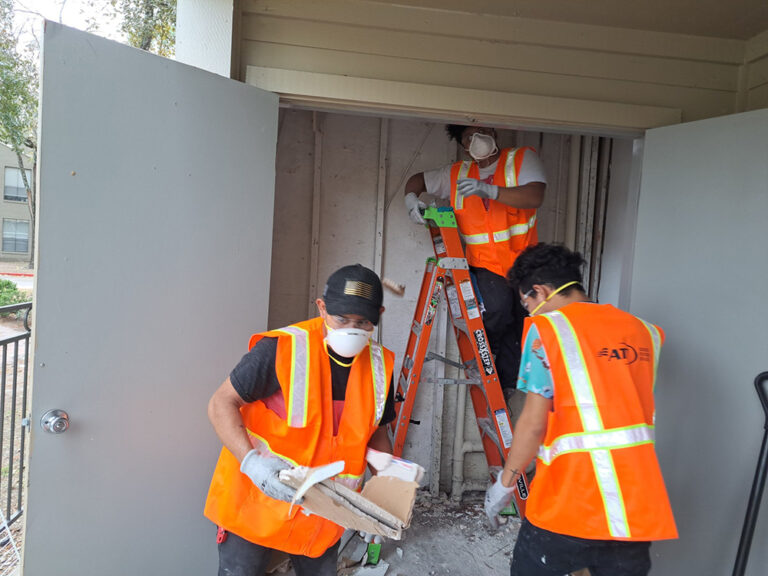 Three workers in safety vests and masks remove debris from a utility closet.