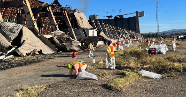 Cleanup crews in protective suits collecting debris from a large burned building.