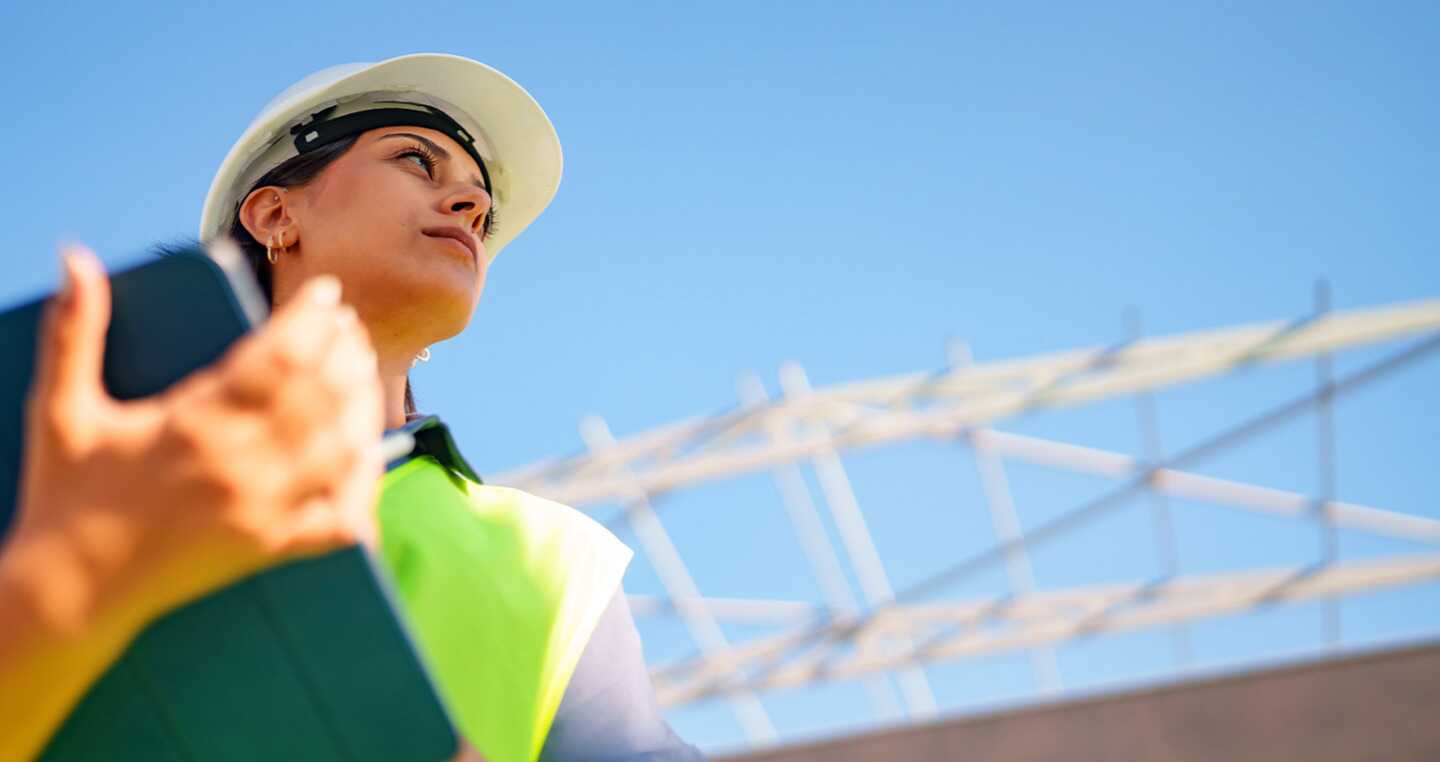 Woman construction worker wearing a hard hat and safety vest, holding a tablet, looks up at a building under construction.