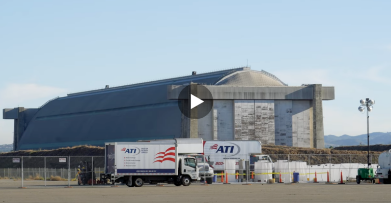 Large, gray, hangar-like structure with two ATI trucks parked in front, along with construction equipment and fencing.